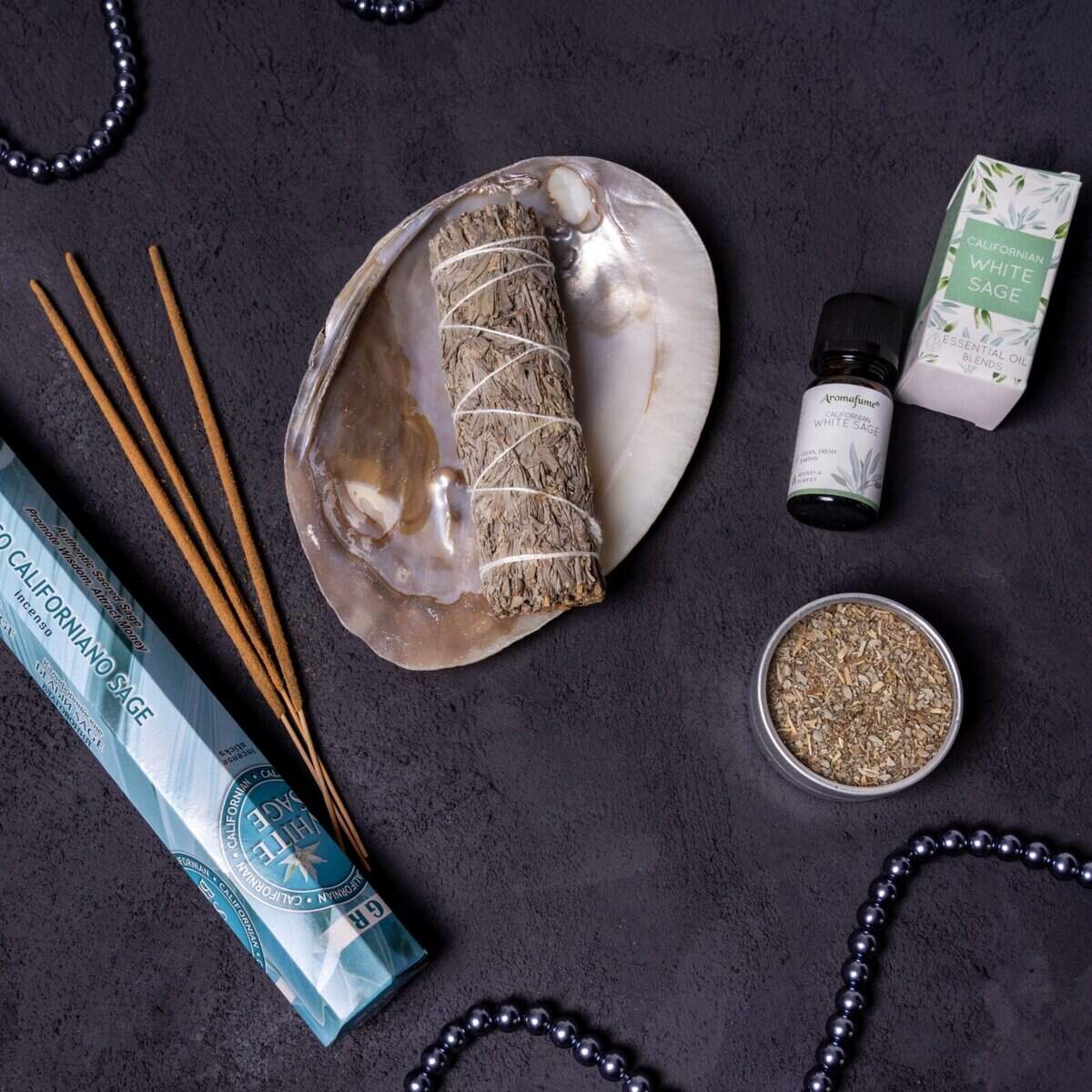 Setul Salvie Albă include un bețișor de salvie într-o coajă, bețișoare de tămâie, ulei esențial, o cutie "Salvie Albă", ierburi uscate într-o cutie și două fire de mărgele negre expuse pe o suprafață întunecată.