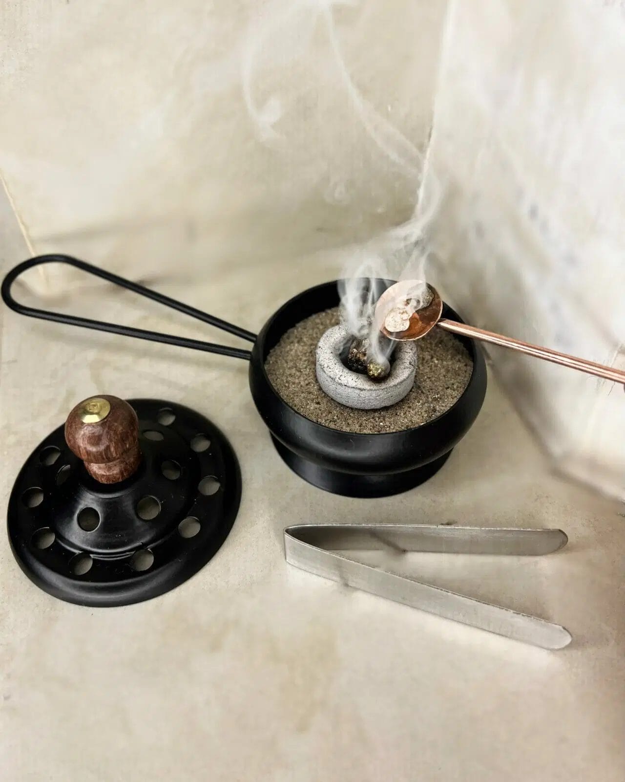 Un arzător de tămâie metalic umplut cu nisip ține un disc de cărbune aprins, din care iese fum. Un capac cu găuri, un clește metalic și o lingură cu rășină sunt așezate în apropiere.